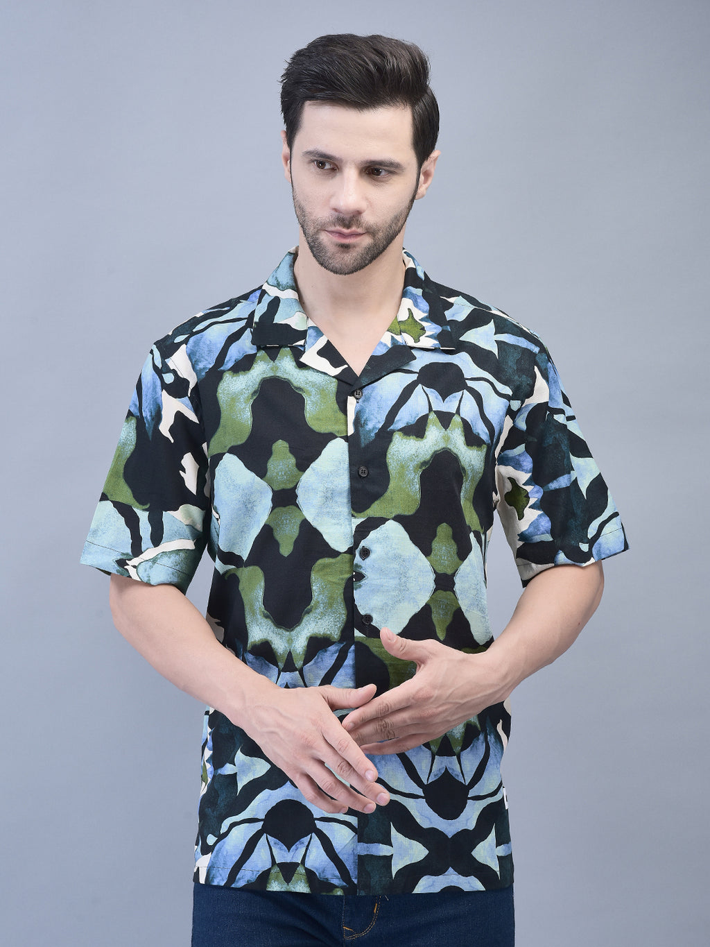 A male model wearing a short-sleeved, geometric abstract print shirt in various colors, paired with blue jeans.