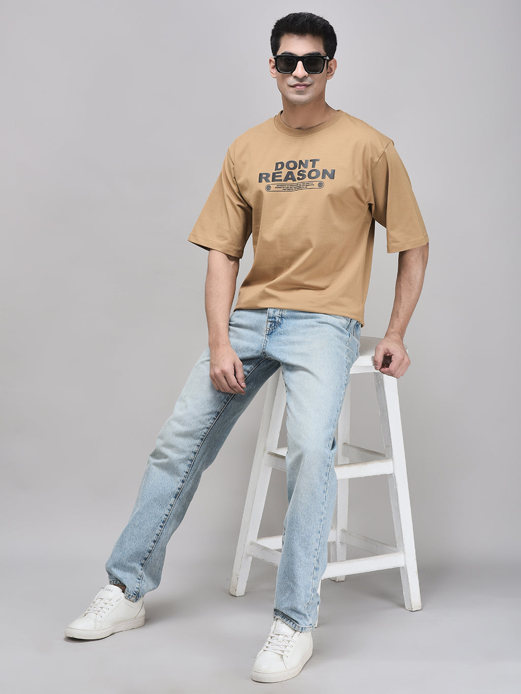 A person wearing a beige 'DON'T REASON' oversized t-shirt with bold lettering graphic, paired with light blue jeans and white sneakers.