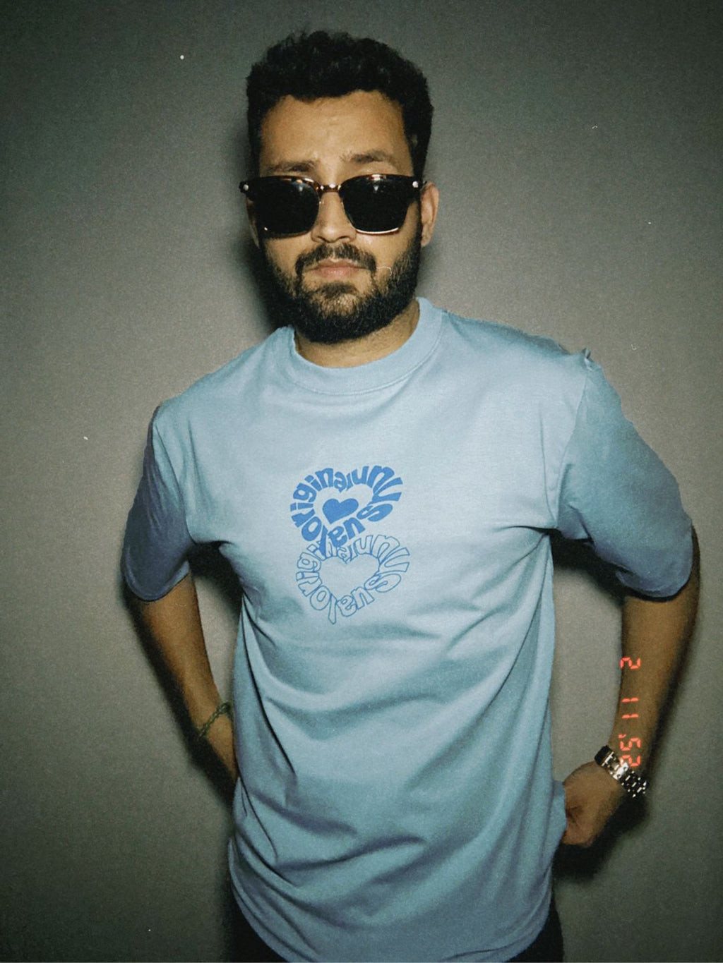 A person wearing a blue oversized t-shirt with text on it, paired with sunglasses and standing against a neutral background.