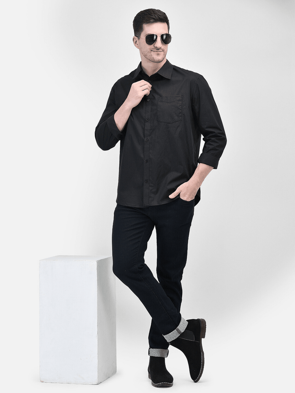 A man wearing a black long-sleeved shirt and black trousers, standing next to a white platform, with his hands in his pockets.