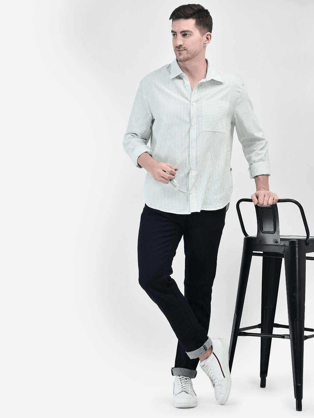 Man wearing green cotton shirt and black pants while standing beside a chair – full front view